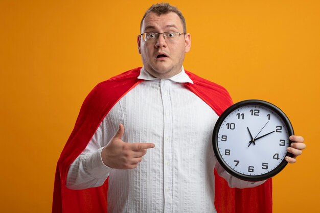 Impresionado hombre superhéroe adulto en capa roja con gafas mirando al frente sosteniendo y apuntando al reloj aislado en la pared naranja