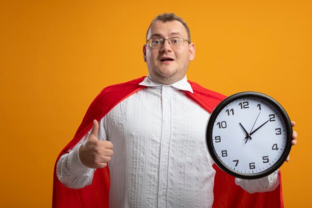 Impresionado hombre superhéroe adulto en capa roja con gafas mirando al frente sosteniendo el reloj mostrando el pulgar hacia arriba aislado en la pared naranja