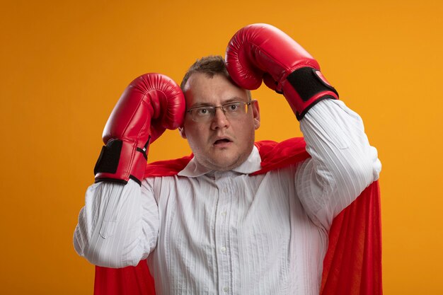 Impresionado hombre superhéroe eslavo adulto en capa roja con gafas y guantes de caja mirando al lado tocando la cabeza con las manos aisladas en la pared naranja