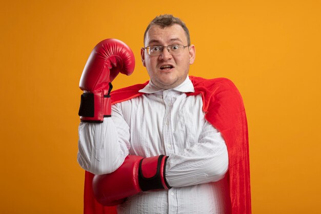 Impresionado hombre superhéroe eslavo adulto en capa roja con gafas y guantes de caja poniendo la mano bajo el brazo manteniendo otra mano en el aire aislada en la pared naranja