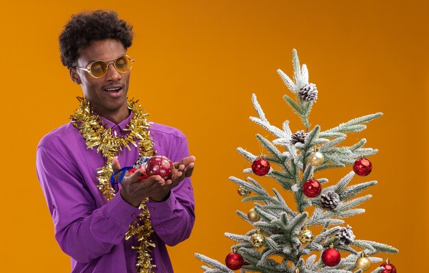 Impresionado joven afroamericano con gafas con guirnalda de oropel alrededor del cuello de pie cerca del árbol de Navidad decorado sosteniendo y mirando adornos navideños aislados sobre fondo naranja