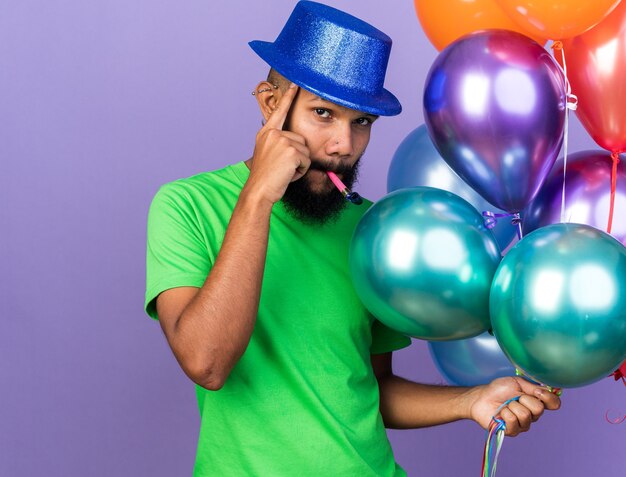 Impresionado joven afroamericano con gorro de fiesta sosteniendo globos soplando silbato de fiesta poniendo el dedo en el templo aislado en la pared azul