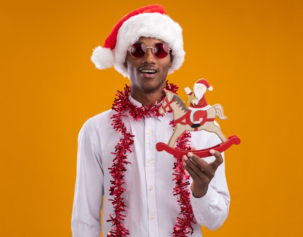 Impresionado joven afroamericano con gorro de Papá Noel y gafas con guirnalda de oropel alrededor del cuello sosteniendo a santa en una figura de caballo mecedora mirando a cámara aislada sobre fondo naranja