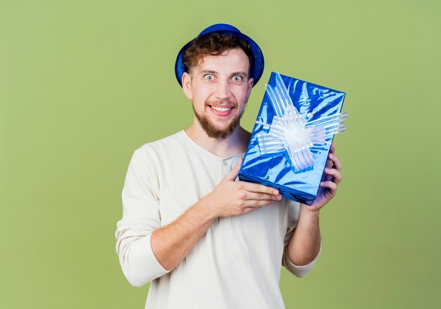 Impresionado joven apuesto chico de fiesta eslava con sombrero de fiesta mostrando caja de regalo mirando a la cámara sonriendo aislado sobre fondo verde oliva con espacio de copia
