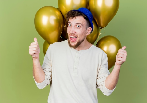 Impresionado joven apuesto chico de fiesta eslavo con sombrero de fiesta de pie delante de globos mirando al frente mostrando los pulgares para arriba aislados en la pared verde oliva