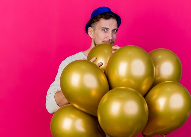 Impresionado joven apuesto chico de fiesta eslavo con sombrero de fiesta de pie detrás de globos sosteniendo uno de ellos mirando a cámara aislada sobre fondo carmesí con espacio de copia