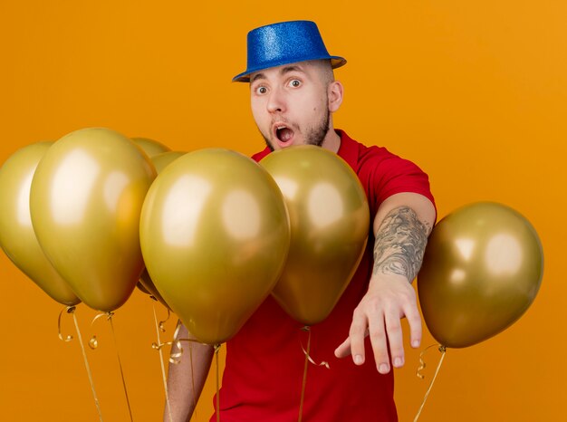 Impresionado joven apuesto chico de fiesta eslavo con sombrero de fiesta de pie entre globos mirando a la cámara estirando la mano hacia la cámara aislada sobre fondo naranja