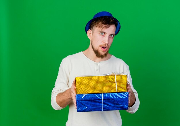 Impresionado joven apuesto chico de fiesta eslavo con sombrero de fiesta sosteniendo cajas de regalo mirando al frente aislado en la pared verde con espacio de copia