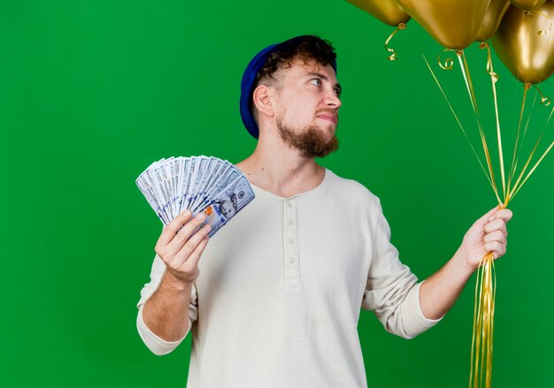 Impresionado joven apuesto chico de fiesta eslavo con sombrero de fiesta sosteniendo globos y dinero aislado sobre fondo verde con espacio de copia