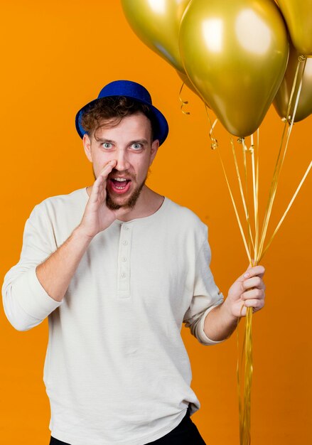 Impresionado joven apuesto chico de fiesta eslavo con sombrero de fiesta sosteniendo globos mirando al frente susurrando aislado en la pared naranja