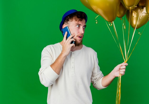 Impresionado joven apuesto chico de fiesta eslavo con sombrero de fiesta sosteniendo globos mirando al lado hablando por teléfono aislado sobre fondo verde con espacio de copia