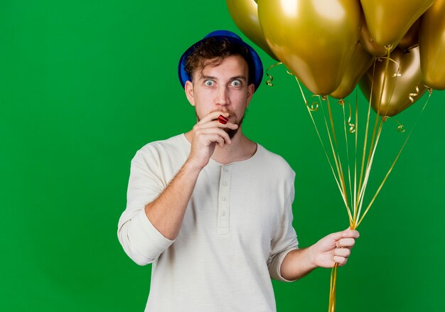 Impresionado joven apuesto chico de fiesta eslavo con sombrero de fiesta sosteniendo globos mirando a cámara poniendo soplador de fiesta en la boca aislada sobre fondo verde con espacio de copia