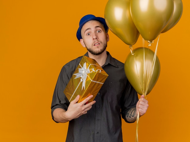 Impresionado joven apuesto chico de fiesta eslavo con sombrero de fiesta sosteniendo globos y paquete de regalo mirando a cámara aislada sobre fondo naranja con espacio de copia