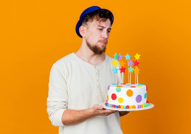 Impresionado joven apuesto chico de fiesta eslavo con sombrero de fiesta sosteniendo pastel de cumpleaños con estrellas mirando directamente aislado sobre fondo naranja con espacio de copia