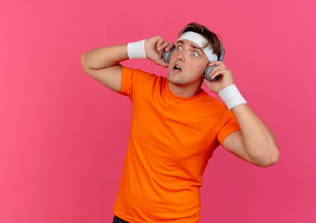 Impresionado joven apuesto deportivo con diadema y muñequeras y auriculares sosteniendo auriculares y mirando al lado aislado en rosa