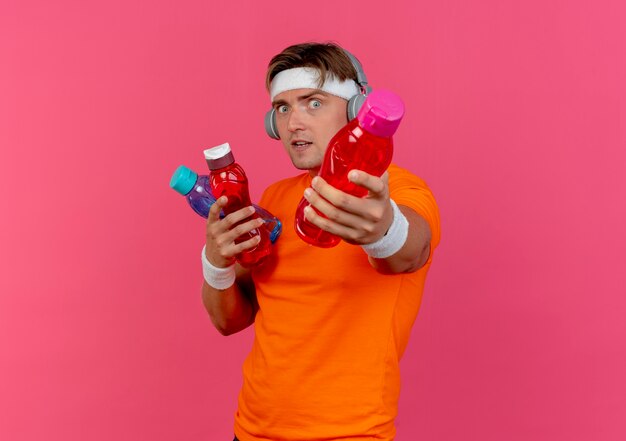 Impresionado joven apuesto deportivo con diadema y muñequeras y auriculares sosteniendo y estirando botellas de agua aisladas en rosa