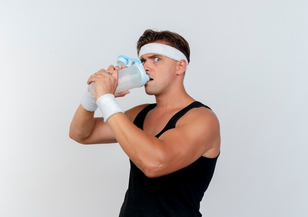 Impresionado joven apuesto deportivo con diadema y muñequeras bebiendo agua de una botella de agua aislada en blanco
