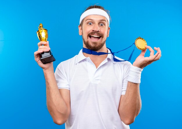 Impresionado joven apuesto deportivo con diadema y muñequeras y medalla alrededor del cuello sosteniendo la medalla y la copa ganadora aislada en la pared azul