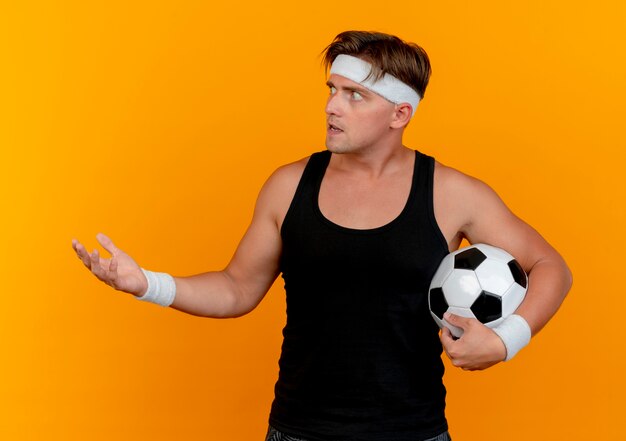 Impresionado joven apuesto deportivo con diadema y muñequeras sosteniendo un balón de fútbol mirando de lado y mostrando la mano vacía aislada en naranja