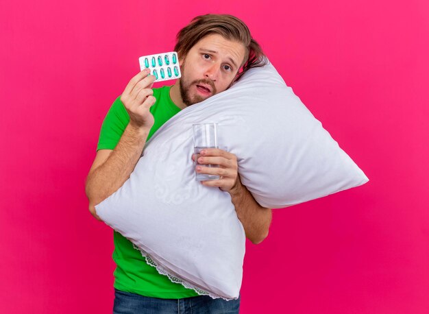 Impresionado joven apuesto enfermo eslavo abrazando la almohada mirando al frente sosteniendo un paquete de cápsulas y un vaso de agua aislado en la pared rosa