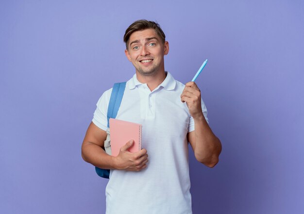 Impresionado joven apuesto estudiante vistiendo bolsa trasera sosteniendo el cuaderno con lápiz aislado en la pared azul