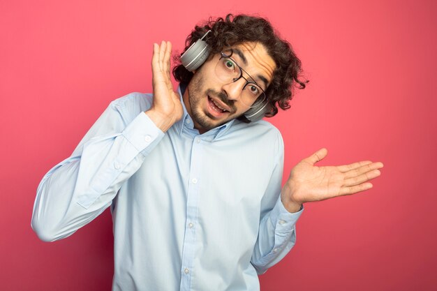 Impresionado joven apuesto hombre caucásico con gafas y auriculares mirando a la cámara manteniendo la mano cerca de la cabeza mostrando la mano vacía aislada sobre fondo carmesí