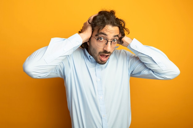 Impresionado joven apuesto hombre caucásico con gafas poniendo las manos en la cabeza mirando a la cámara aislada sobre fondo naranja