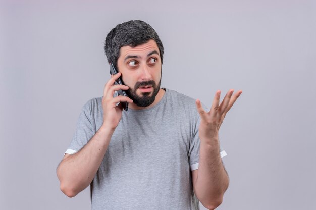 Impresionado joven apuesto hombre caucásico hablando por teléfono mirando de lado y mostrando la mano vacía aislada en blanco con espacio de copia