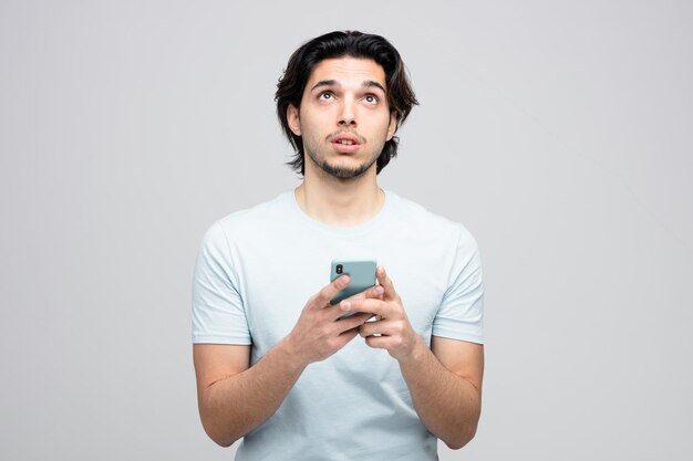 Impresionado joven apuesto hombre sosteniendo el teléfono móvil con ambas manos mirando hacia arriba aislado sobre fondo blanco.