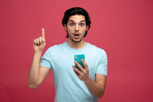 Impresionado joven apuesto hombre sosteniendo teléfono móvil mirando a la cámara apuntando hacia arriba aislado sobre fondo rojo.
