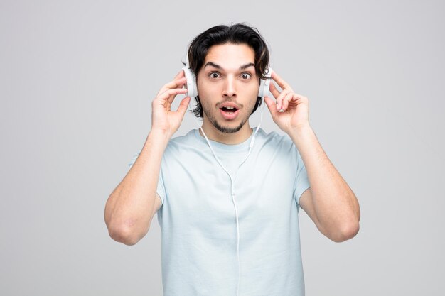 Impresionado joven apuesto hombre usando y tocando auriculares mirando a la cámara aislada sobre fondo blanco.