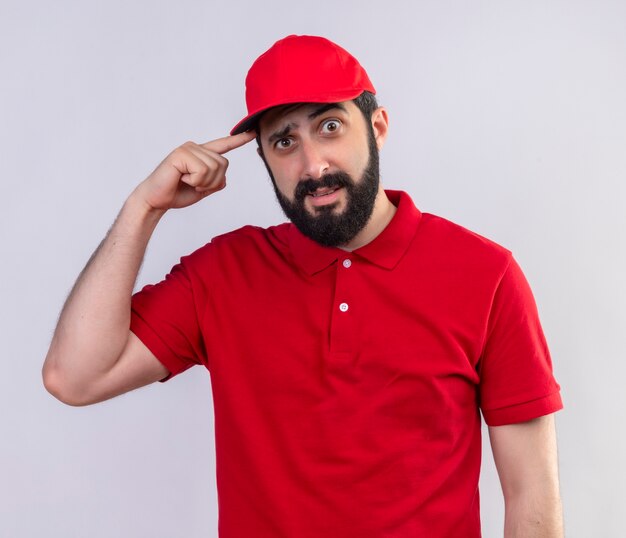 Impresionado joven apuesto repartidor caucásico con uniforme rojo y gorra poniendo el dedo en la sien aislado en blanco