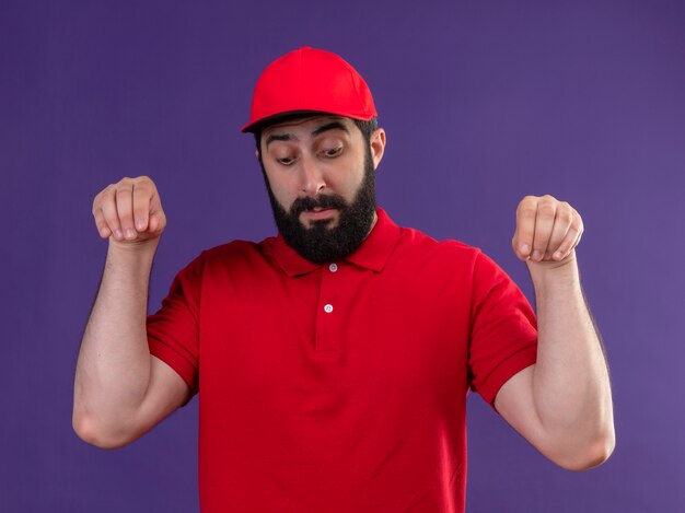 Impresionado joven apuesto repartidor caucásico vestido con uniforme rojo y gorra fingir sosteniendo algo mirando hacia abajo aislado en púrpura