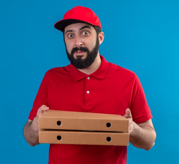 Impresionado joven apuesto repartidor caucásico vestido con uniforme rojo y gorra sosteniendo cajas de pizza aisladas en azul