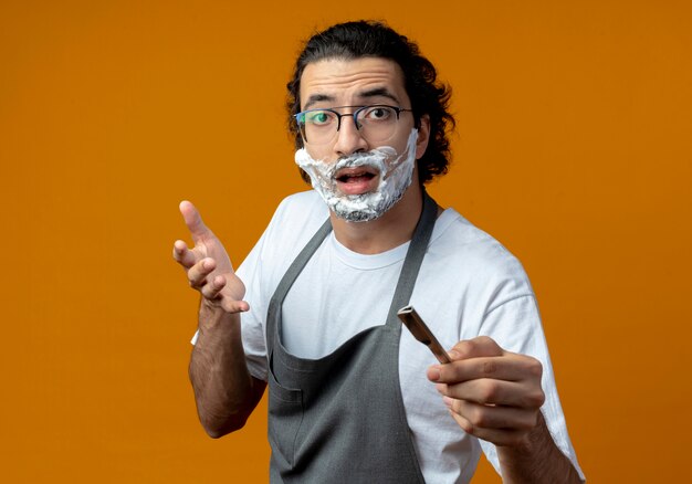 Impresionado joven barbero caucásico con gafas y banda para el pelo ondulado en uniforme sosteniendo una navaja de afeitar con crema de afeitar puesta en su rostro sosteniendo una navaja de afeitar y manteniendo la mano en el aire