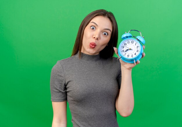 Impresionado joven bonita mujer sosteniendo despertador aislado en verde con espacio de copia