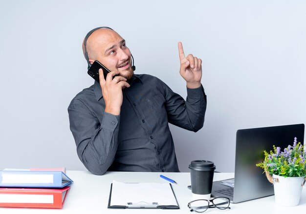 Impresionado joven calvo call center hombre con auriculares sentado en el escritorio con herramientas de trabajo hablando por teléfono levantando el dedo y mirando al lado aislado en blanco