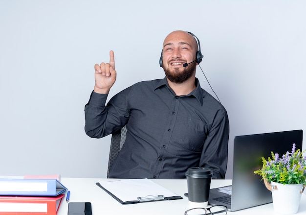 Impresionado joven calvo call center hombre con auriculares sentado en el escritorio con herramientas de trabajo mirando y apuntando hacia arriba aislado en blanco