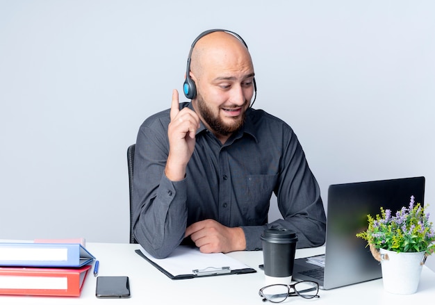 Impresionado joven calvo call center hombre con auriculares sentado en el escritorio con herramientas de trabajo mirando portátil y levantando el dedo aislado en blanco