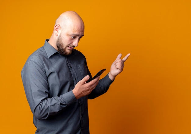 Foto gratuita impresionado joven calvo call center hombre de pie en la vista de perfil sosteniendo y mirando el teléfono móvil y apuntando al lado aislado en naranja con espacio de copia