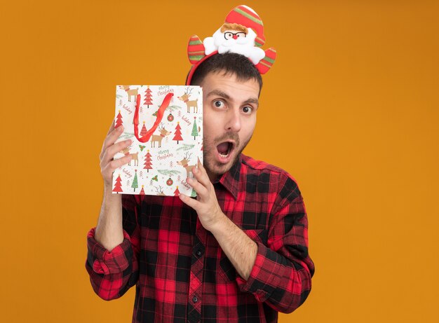 Impresionado joven caucásico con diadema de santa claus con bolsa de regalo de navidad cerca de la cabeza aislada en la pared naranja con espacio de copia