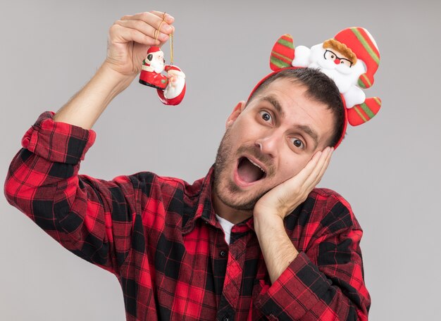 Impresionado joven caucásico con diadema de santa claus levantando adornos navideños de santa claus mirando a cámara manteniendo la mano en la cara aislada sobre fondo blanco