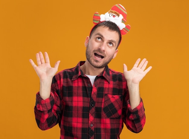 Impresionado joven caucásico con diadema de santa claus mostrando las manos vacías mirando hacia arriba aislado en la pared naranja