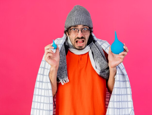 Impresionado joven caucásico enfermo con gafas, gorro de invierno y bufanda envuelto en cuadros con enemas mirando a cámara aislada sobre fondo carmesí con espacio de copia