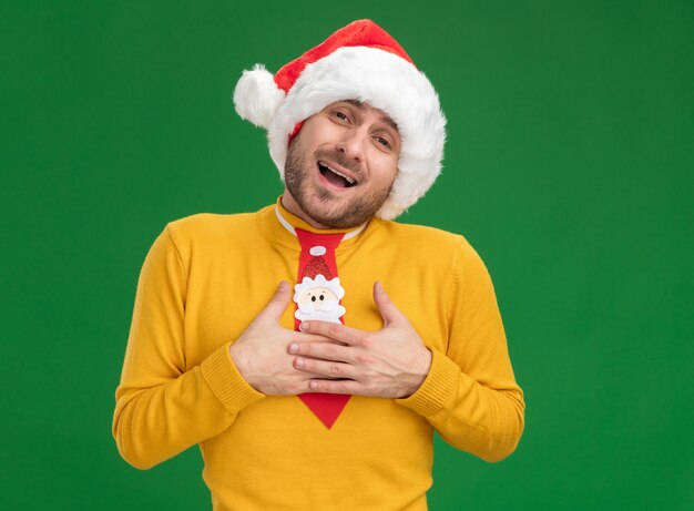 Impresionado joven caucásico con sombrero de navidad y corbata haciendo gesto de agradecimiento aislado en la pared verde con espacio de copia