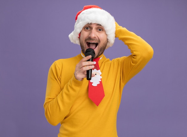 Impresionado joven caucásico con sombrero de navidad y corbata sosteniendo el micrófono cerca de la boca mirando a la cámara manteniendo la mano en la cabeza aislada sobre fondo púrpura