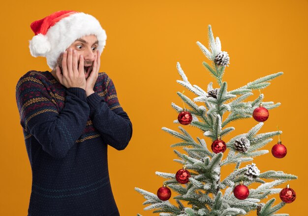 Impresionado joven caucásico con sombrero de navidad de pie cerca del árbol de navidad decorado manteniendo las manos en la cara mirando el árbol aislado en la pared naranja