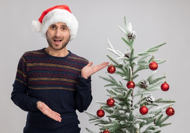 Impresionado joven caucásico con sombrero de navidad de pie cerca del árbol de navidad mirando a la cámara apuntando con las manos al árbol aislado sobre fondo blanco.