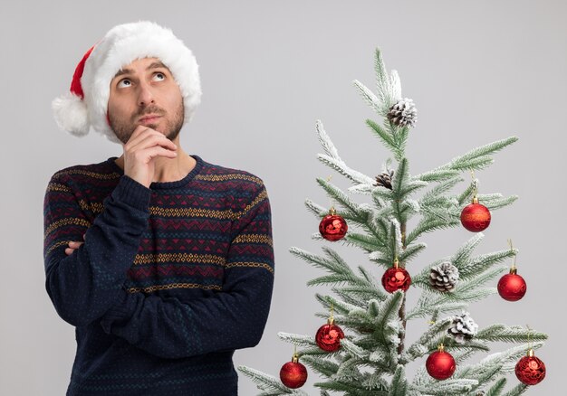 Impresionado joven caucásico con sombrero de navidad de pie cerca del árbol de navidad tocando la barbilla mirando hacia arriba aislado en la pared blanca