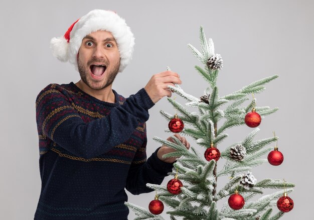 Impresionado joven caucásico con sombrero de navidad de pie cerca del árbol de navidad tocándolo aislado en la pared blanca